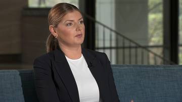 Kobieta w ciemnej marynarce i białej koszuli m&oacute;wi, siedząc na kanapie. W tle widać schody i okna.