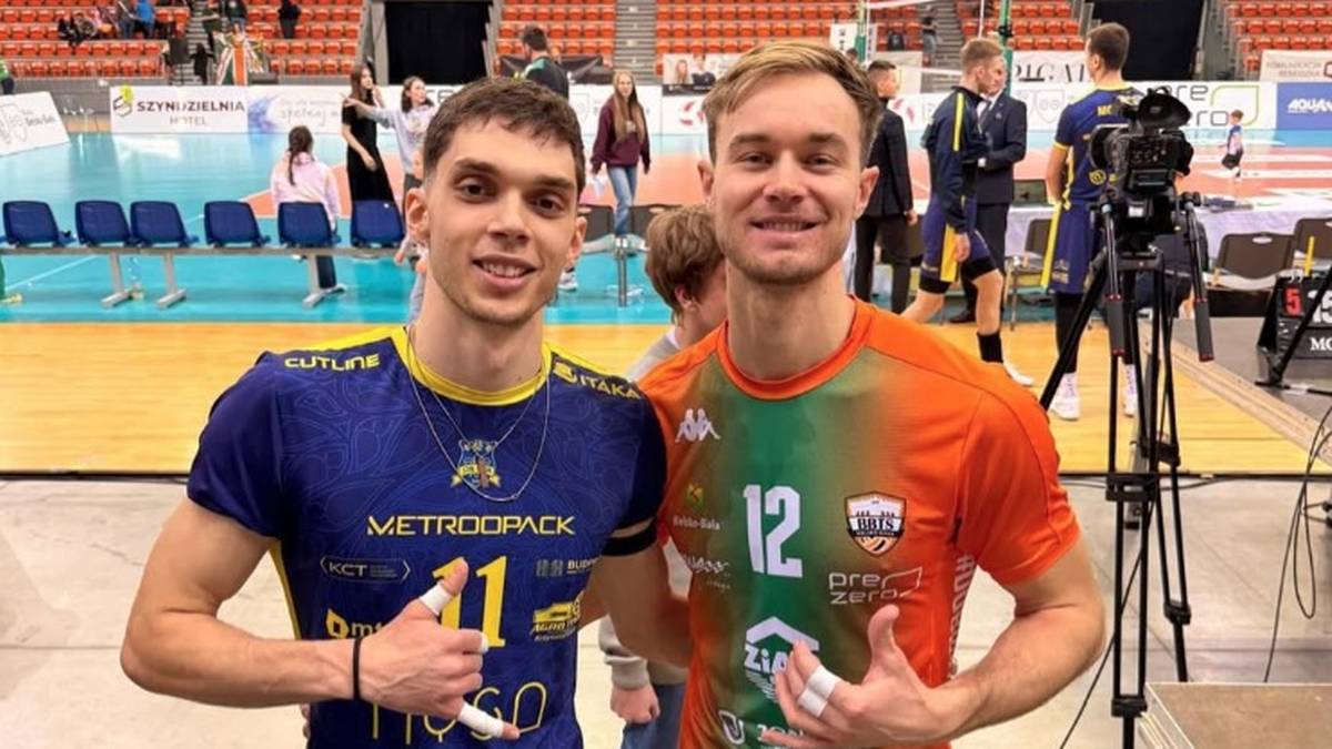 Dw&oacute;ch młodych mężczyzn w strojach sportowych, prawdopodobnie siatkarzy, pozuje do zdjęcia w hali sportowej. Obaj uśmiechają się do aparatu.