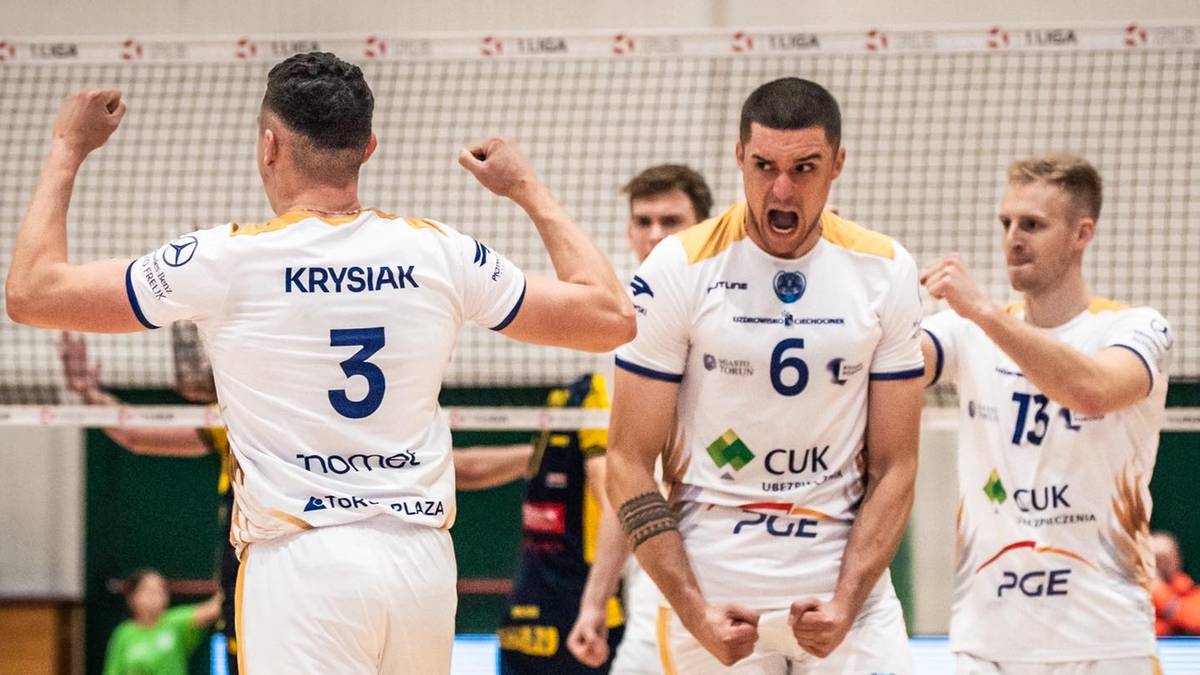 Trzech siatkarzy w białych strojach z numerami na plecach unosi ręce w geście radości, krzycząc w stronę siatki.