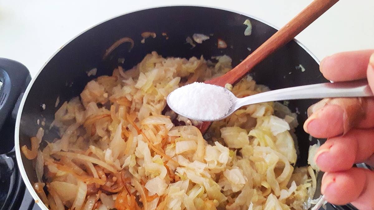 Zbliżenie na garnek z gotującą się kapustą, do której z łyżeczki dodawana jest sól.