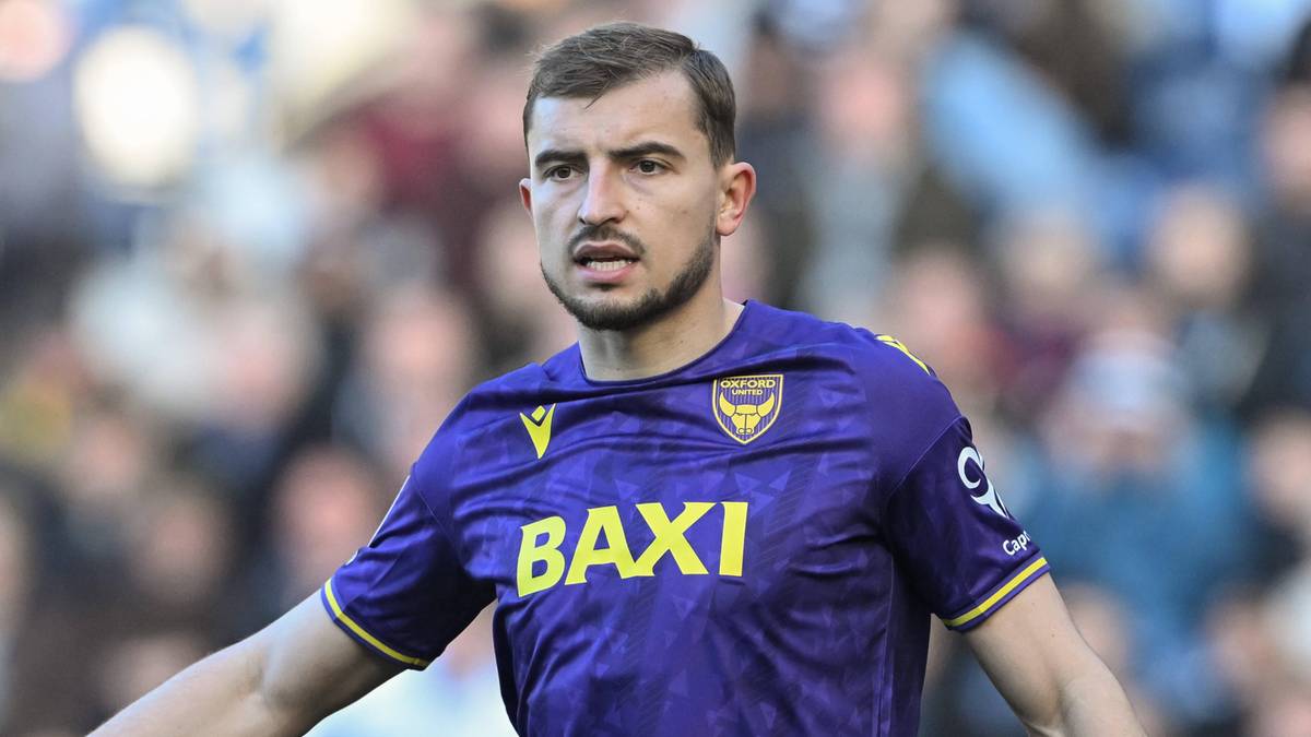 Młody mężczyzna o ciemnych włosach i z zarostem, ubrany w fioletową koszulkę piłkarską z żółtym napisem "BAXI" i herbem klubu na piersi, stoi na boisku piłkarskim podczas meczu.