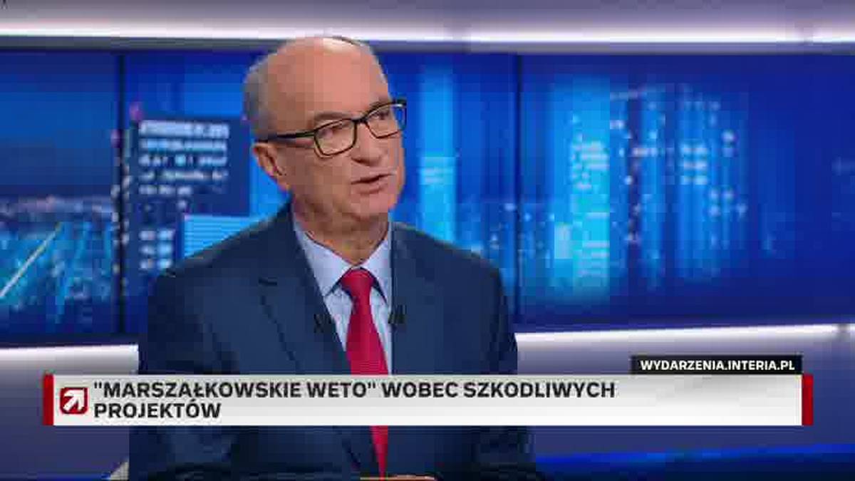 Mężczyzna w garniturze i czerwonym krawacie mówi do kamery. W tle znajduje się podświetlany ekran z miejskim pejzażem nocą i banerem informacyjnym.