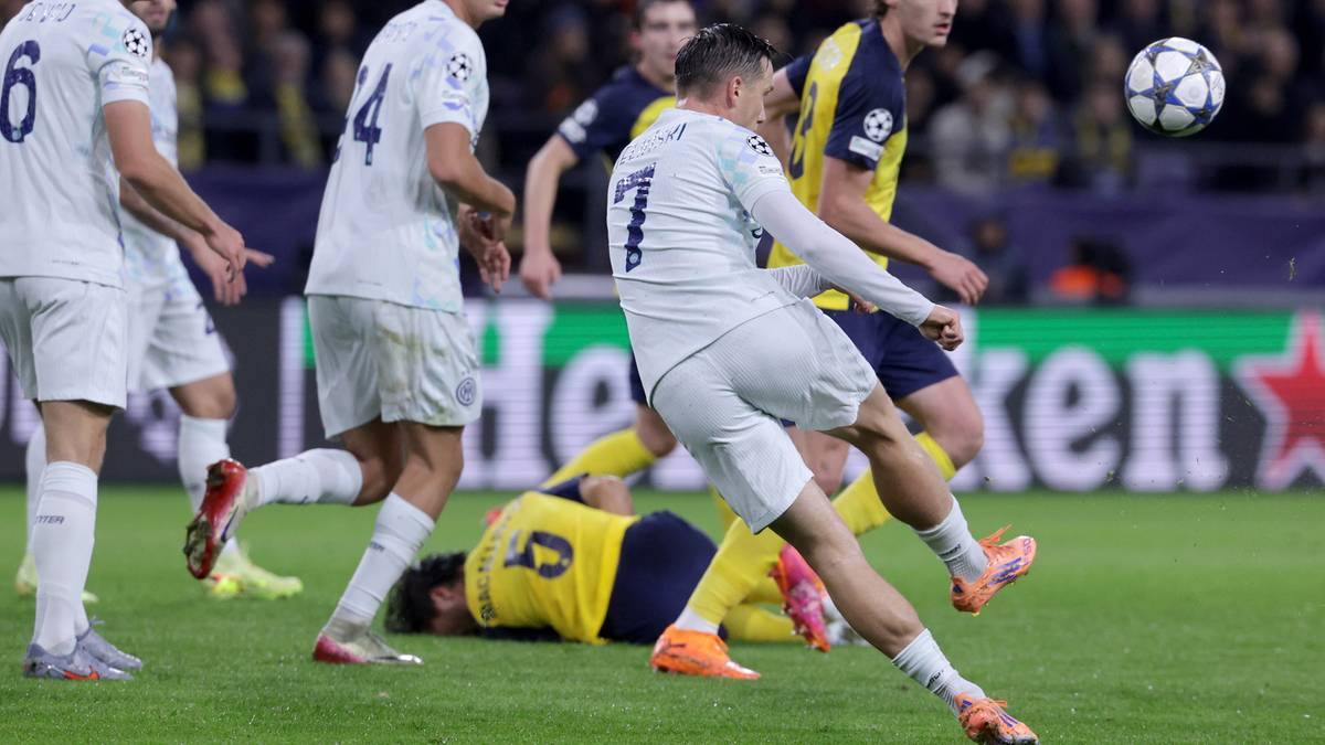 Piłkarz w białej koszulce wykonuje strzał na bramkę podczas meczu piłki nożnej. W tle widać innych zawodników i fragment trybun.