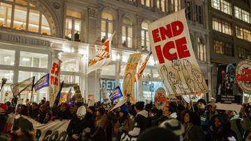 Tłum ludzi protestujących na ulicy nocą, trzymający transparenty z hasłami, w tym jeden z napisem "END ICE". W tle budynek z oświetlonymi oknami.