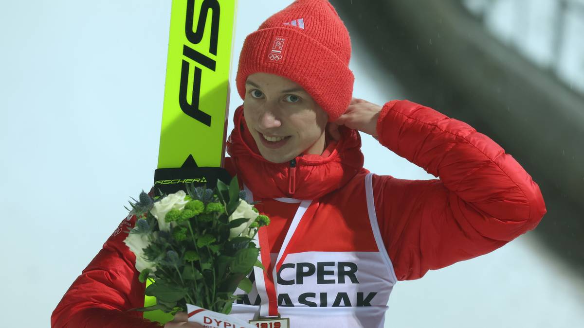 Skoczek narciarski w czerwonym stroju i czapce, trzymający bukiet białych kwiat&oacute;w i medal na szyi, z nartą FIS w tle.