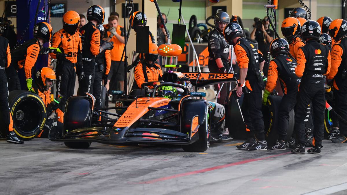 Zespół mechaników w pomarańczowych strojach otacza bolid Formuły 1 w boksie.