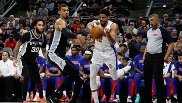 Gracz w białym stroju z piłką w rękach, otoczony przez przeciwnik&oacute;w podczas meczu koszyk&oacute;wki.