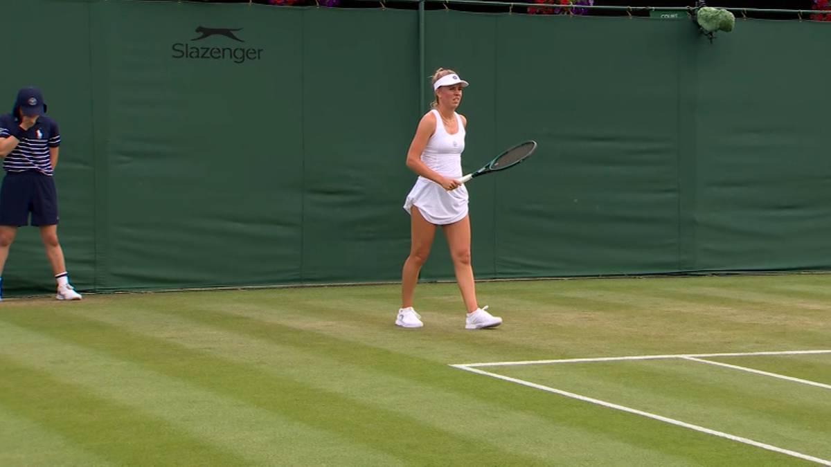 Tenisistka Magdalena Fręch w białej sukience i czapce z daszkiem porusza się po korcie tenisowym z rakietą w dłoni. Na drugim planie widoczna jest inna osoba w granatowej koszulce i spodenkach.