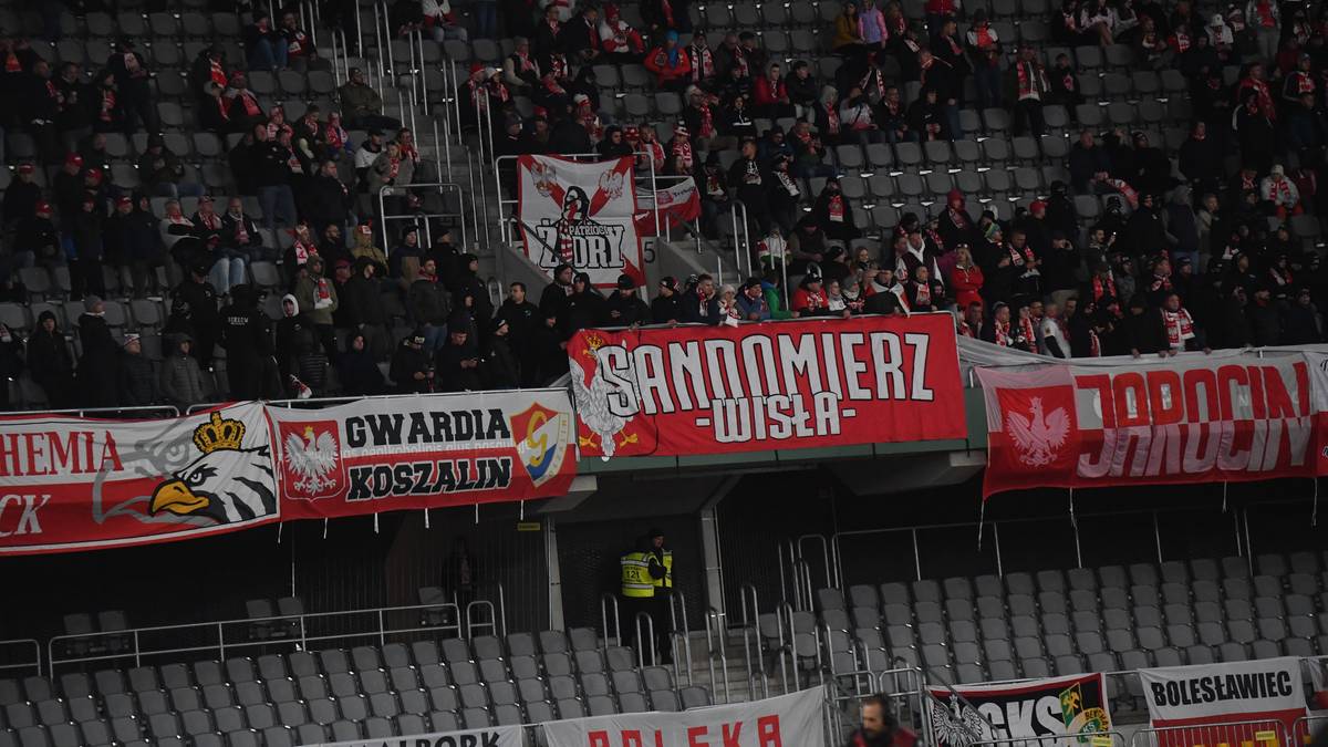 Tłum kibiców na trybunach stadionu, widoczne transparenty z nazwami grup kibicowskich i hasłami.