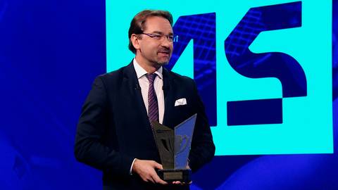 Mężczyzna w garniturze odbiera nagrodę na tle niebieskiego ekranu z cyframi "15".