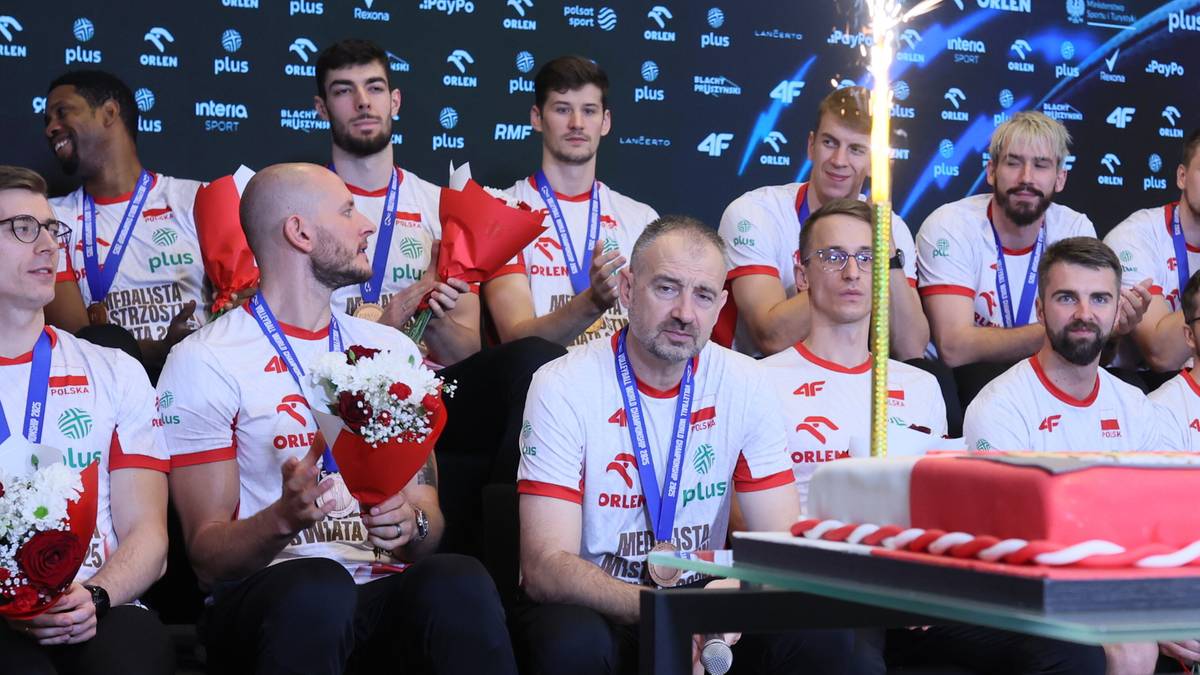 Grupa siatkarzy w strojach reprezentacyjnych i z medalami na szyjach, siedząca na podium. Na pierwszym planie kilku zawodników, a z tyłu trener Nikola Grbić i inni członkowie zespołu. W tle widać banery sponsorów, a na stole przed zawodnikami tort i race.