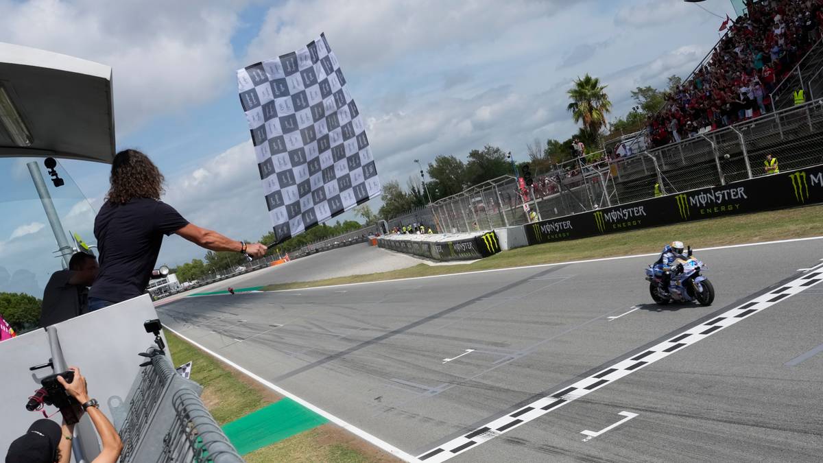 Motocyklista zbliża się do mety, a osoba po lewej stronie macha flagą w szachownicę.