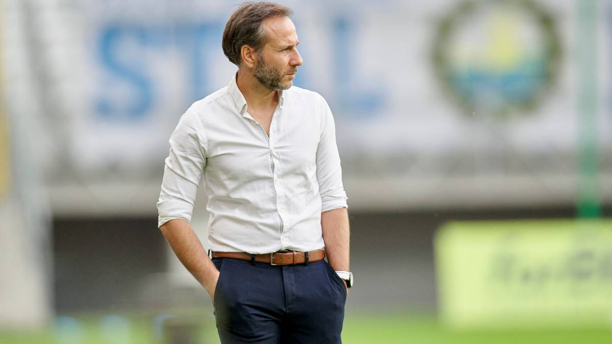 Mężczyzna w białej koszuli z podwiniętymi rękawami i granatowych spodniach stoi na boisku piłkarskim, patrząc w bok z rękami w kieszeniach.