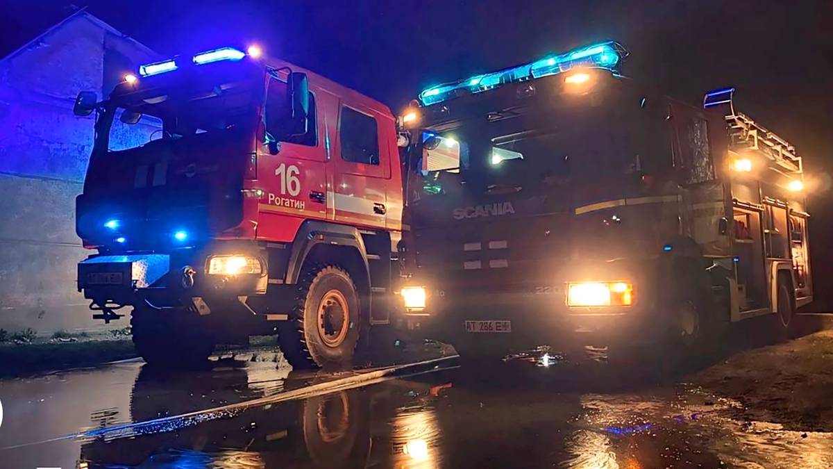 Dwa wozy strażackie stojące nocą na mokrej nawierzchni, oświetlone niebieskimi i żółtymi światłami.