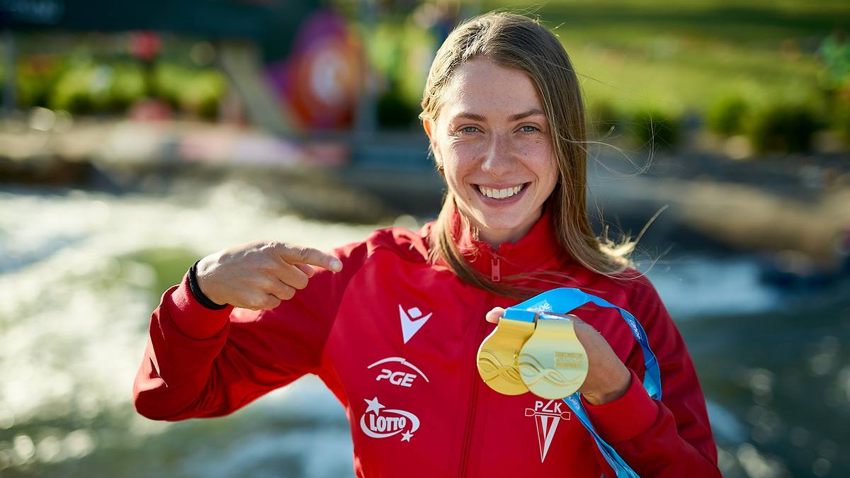 Klaudia Zwolińska, polska kajakarka, w czerwonej bluzie sportowej z dwoma złotymi medalami na smyczach, radośnie uśmiecha się do kamery, wskazując na swoje osiągnięcie.