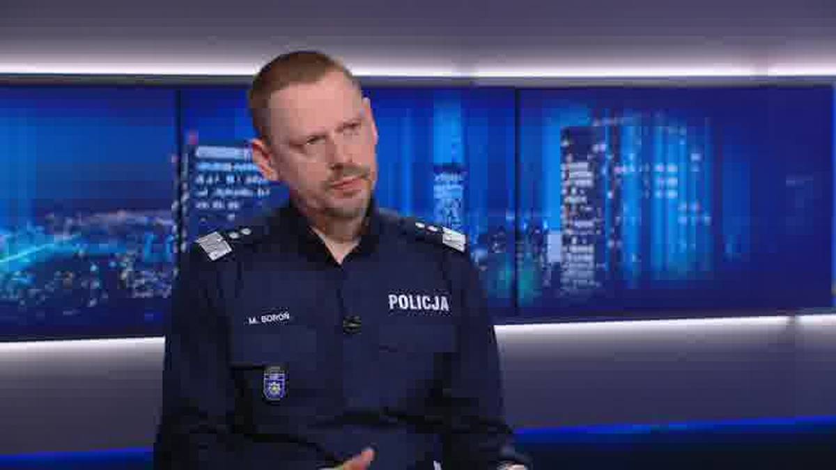 Mężczyzna w mundurze policji z naszywką "POLICJA" na piersi, siedzi na tle ekran&oacute;w z nocną panoramą miasta.