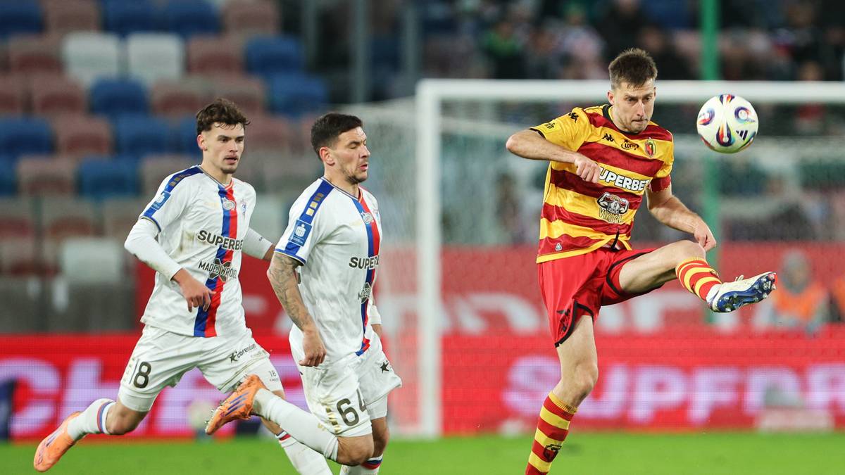 Piłkarz w żółto-czerwonym stroju odbija piłkę nogą podczas meczu, w tle dwaj zawodnicy w białych strojach.