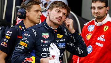 Max Verstappen, kierowca Formuły 1, w słuchawkach i kombinezonie, poprawia słuchawkę w uchu. W tle widać innych członk&oacute;w zespoł&oacute;w, w tym osoby w strojach zespoł&oacute;w Red Bull i Ferrari.