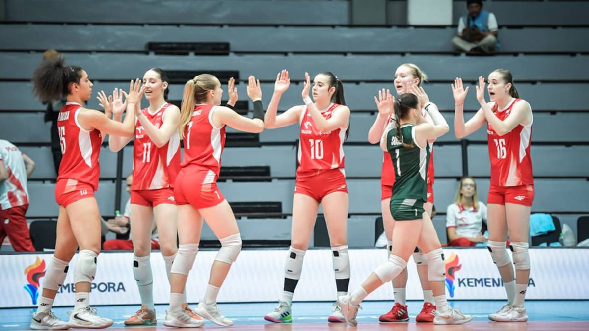 Dwie drużyny siatkarek po meczu wykonują sobie "piątki". Zawodniczki w czerwonych strojach są po lewej stronie, a w zielonych po prawej.