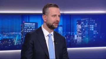 Mężczyzna w garniturze i krawacie m&oacute;wi, na tle panelu z widokiem nocnego miasta.