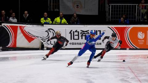 Trzech łyżwiarzy szybkich rywalizuje na lodzie. Zawodnik w niebieskim stroju prowadzi, a za nim jadą zawodnicy w czerwonym i czarnym stroju.