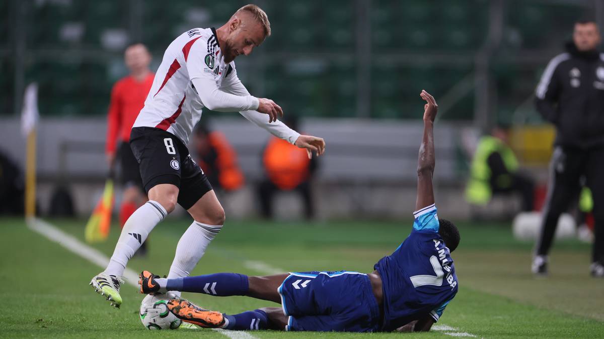 Dwóch piłkarzy podczas meczu. Jeden w białej koszulce z numerem 8 próbuje odebrać piłkę leżącemu na ziemi zawodnikowi w niebieskim stroju.