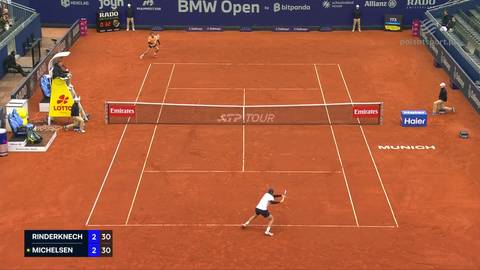 Widok z g&oacute;ry na kort tenisowy z czerwonej mączki. Dw&oacute;ch graczy jest w trakcie meczu. Na dole ekranu znajduje się tablica wynik&oacute;w pokazująca "RINDERKNECH" i "MICHELSEN" z wynikiem 2:30.