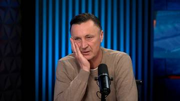 Tomasz Hajto siedzi przy stole, opierając policzek na dłoni, z zamyślonym wyrazem twarzy.