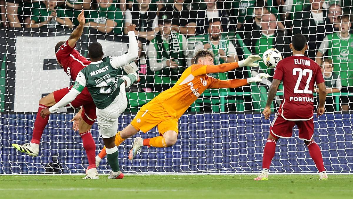 Bramkarz w pomarańczowym stroju broni strzał, obrońca w zielonym stroju próbuje odebrać piłkę, inny zawodnik w czerwonym stroju obserwuje sytuację.