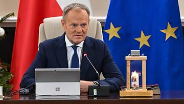 Donald Tusk siedzi przy stole, przed nim tablet i mikrofon, w tle flagi Polski i Unii Europejskiej.