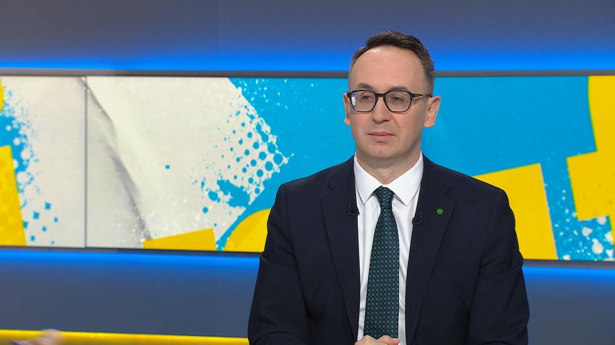 Mężczyzna w garniturze i krawacie, w okularach, siedzi przed kolorowym tłem w studiu telewizyjnym.