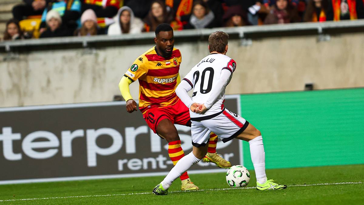 Piłkarz w żółto-czerwonym stroju próbuje odebrać piłkę zawodnikowi w białym stroju z numerem 20.