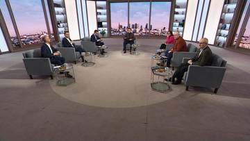 Siedem os&oacute;b siedzi w okręgu na kanapach w studiu telewizyjnym, dyskutując.