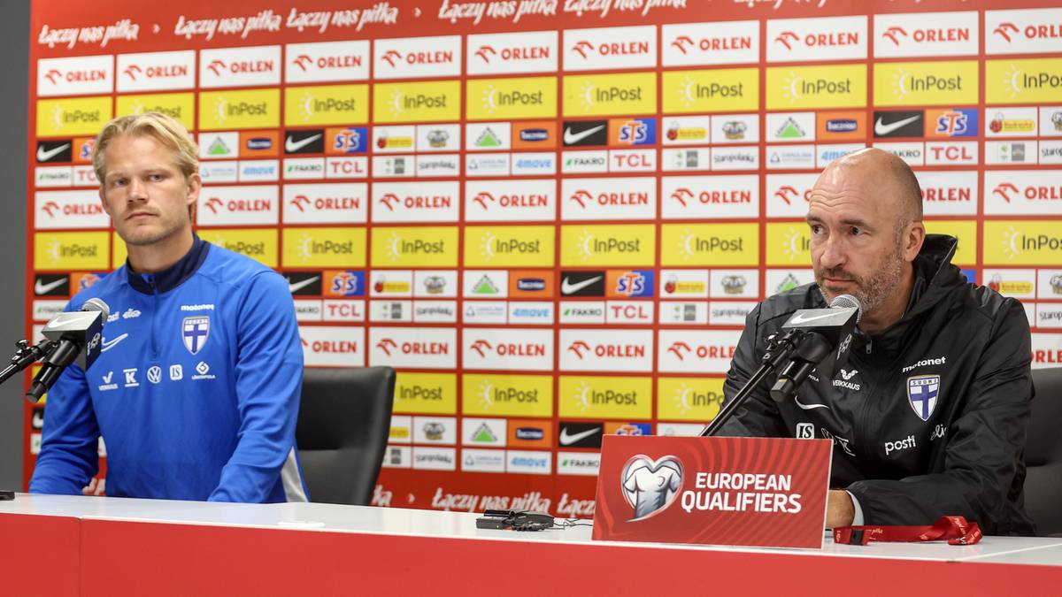 Dwóch mężczyzn siedzi za stołem przed ścianą z logo sponsorów. Po lewej młodszy mężczyzna w niebieskiej bluzie, po prawej starszy mężczyzna w czarnej kurtce, mówiący do mikrofonu. Na stole leży czerwona tabliczka "EUROPEAN QUALIFIERS".