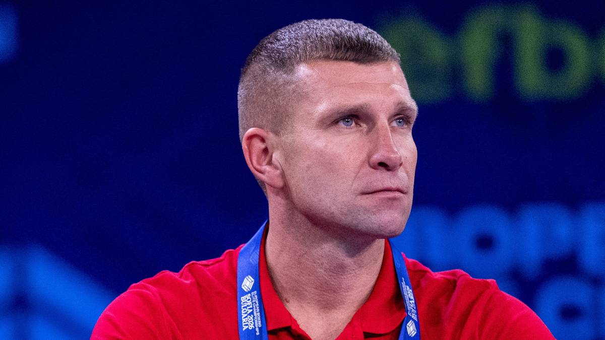 Portret mężczyzny z kr&oacute;tkimi, jasnymi włosami i niebieskimi oczami, ubranego w czerwoną koszulkę polo. Tło jest niebieskie z widocznymi białymi napisami.