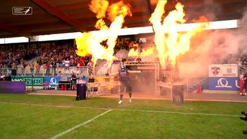 Zawodnik w stroju sportowym wchodzi na boisko, a po obu stronach towarzyszą mu wysokie płomienie ognia i kłęby dymu.
