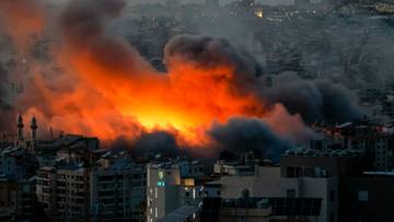 Dynamiczny obraz przedstawiający gęsty, ciemny dym unoszący się nad płonącymi budynkami w mieście nocą. Intensywne, pomarańczowe płomienie przebijają się przez dym, tworząc dramatyczny kontrast z otaczającym mrokiem.