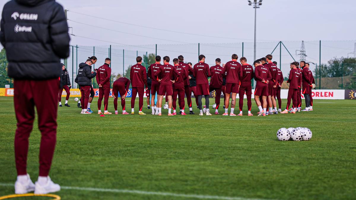 Drużyna piłkarzy w czerwonych strojach stoi na murawie stadionu, w tle widać ludzi w kurtkach i ogrodzenie.