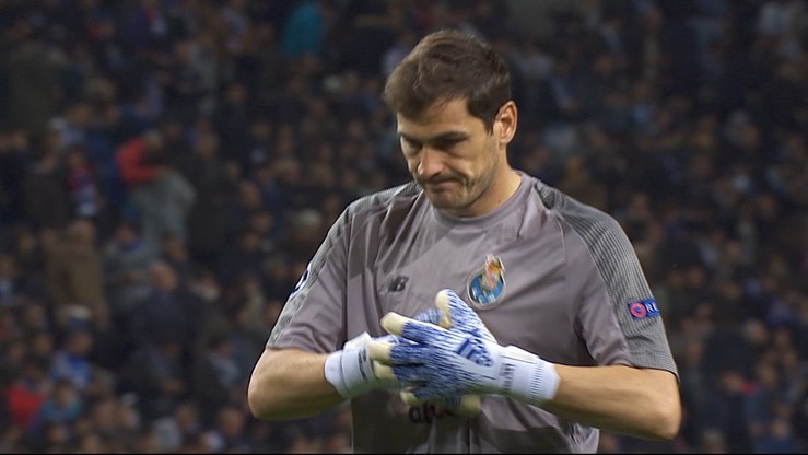 Bramkarz Iker Casillas w niebiesko-szarym stroju bramkarskim, ze złotymi rękawicami bramkarskimi na rękach, stoi na boisku piłkarskim, na tle trybun wypełnionych kibicami.