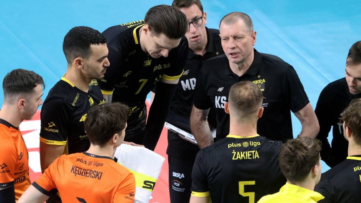 Grupa siatkarzy w czarnych i pomarańczowych strojach, skupionych wok&oacute;ł trenera.