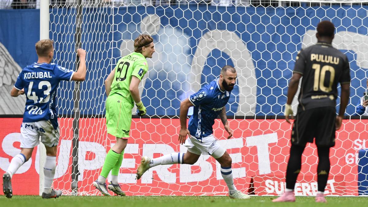 Czterech piłkarzy na boisku piłkarskim przed bramką. Dwóch piłkarzy w niebieskich strojach, jeden w zielonym i jeden w czarnym. Na jednym z niebieskich strojów widoczny numer 43.