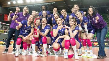 Drużyna siatkarek w granatowo-fioletowych strojach pozuje do wsp&oacute;lnego zdjęcia, trzymając się za ręce i uśmiechając.