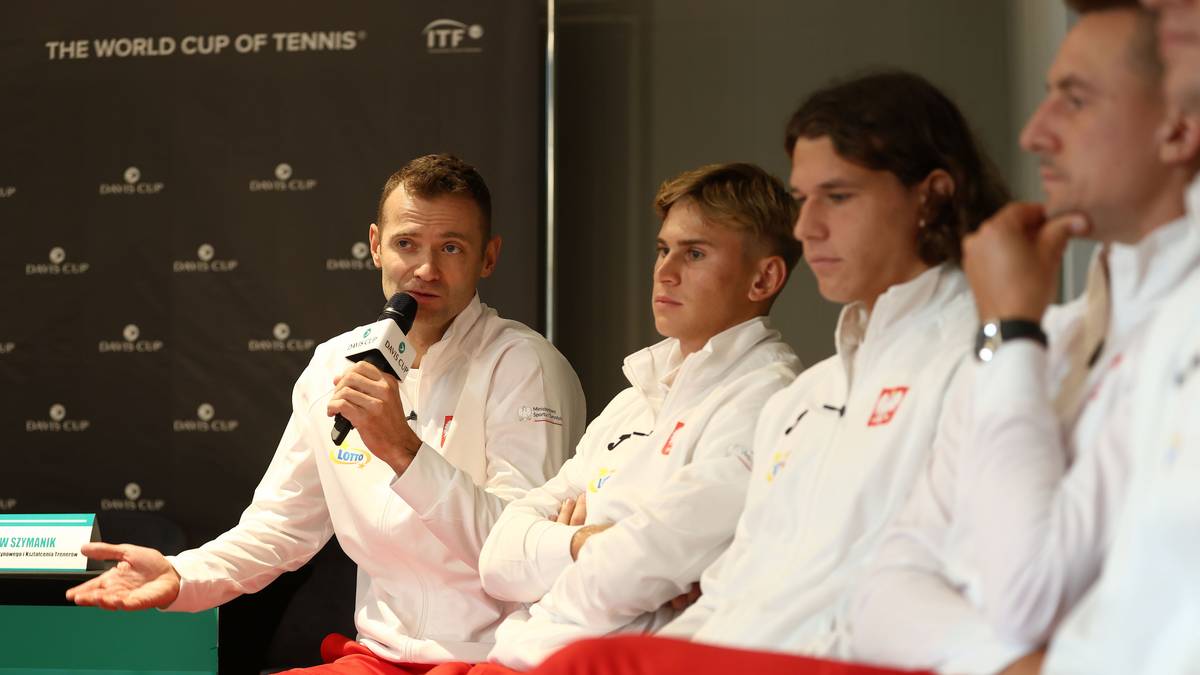 Czterech mężczyzn w białych kurtkach tenisowych siedzi w rzędzie. Jeden z nich, po lewej, mówi do mikrofonu, gestykulując prawą ręką. Pozostali słuchają.