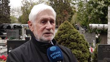 Portret starszego mężczyzny z siwymi włosami i brodą, mówiącego do mikrofonu z logo Polsat Sport. W tle widoczne są nagrobki i drzewa.