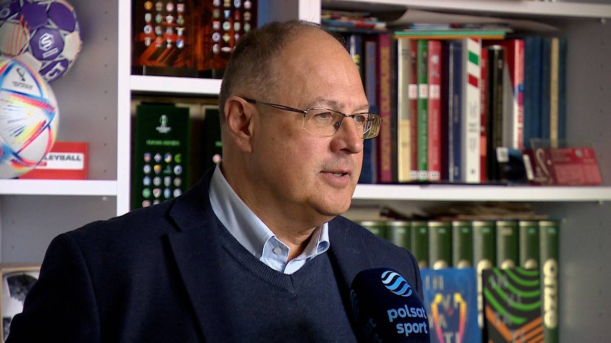 Mężczyzna w okularach i granatowej marynarce, trzymający mikrofon z logo Polsat Sport.