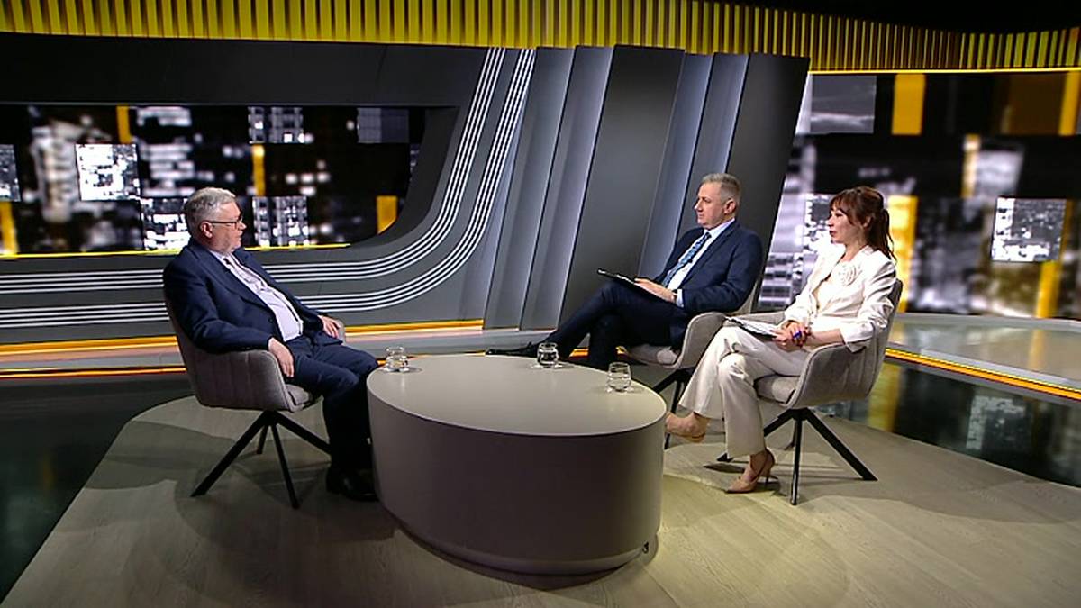 Trzy osoby siedzą w studio telewizyjnym wok&oacute;ł owalnego stołu. Dwie siedzą po lewej stronie, jedna po prawej. Wszyscy siedzą na krzesłach, skierowani do przodu, w kierunku widza. W tle widać elementy scenografii studyjnej z ekranami wyświetlającymi obrazy budynk&oacute;w.