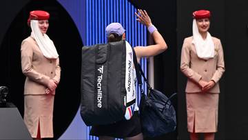 Tenisistka Iga Świątek z torbą sportową między dwiema kobietami w uniformach, z napisem MELBOURNE w tle.