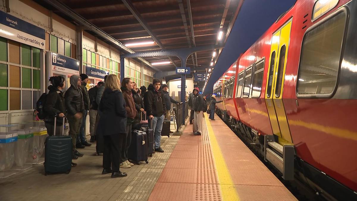 Grupa os&oacute;b stoi na peronie kolejowym obok zaparkowanego czerwonego pociągu, niekt&oacute;rzy z nich mają bagaże.