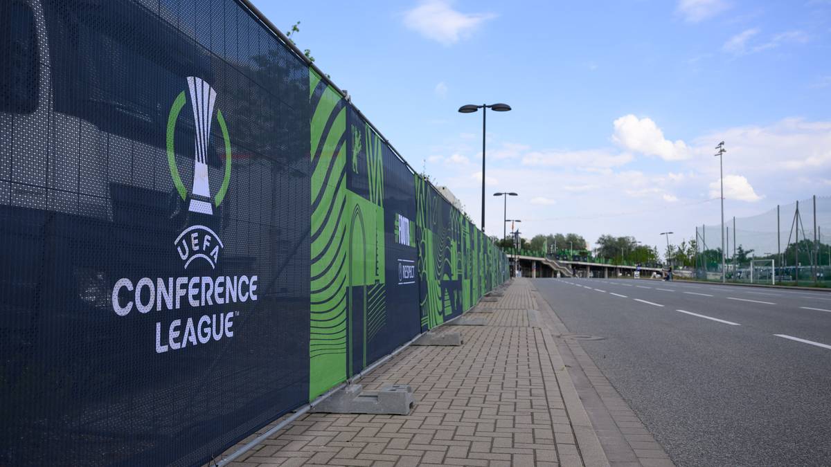 Bariera ogrodzeniowa z logo Ligi Konferencji UEFA i napisem "CONFERENCE LEAGUE" w kolorze białym na ciemnym tle. Obok znajduje się droga i chodnik.