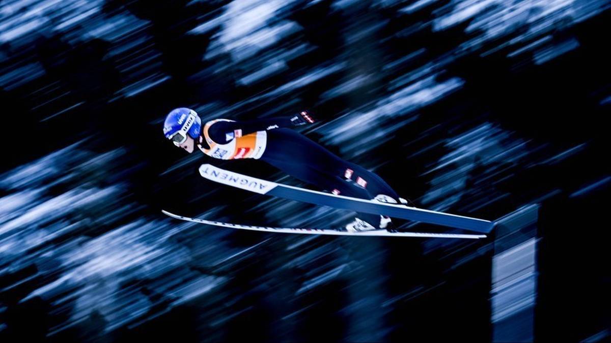 Skoczek narciarski w trakcie lotu na nartach. Zawodnik ubrany jest w ciemny kombinezon z pomarańczowymi elementami i niebieski kask.